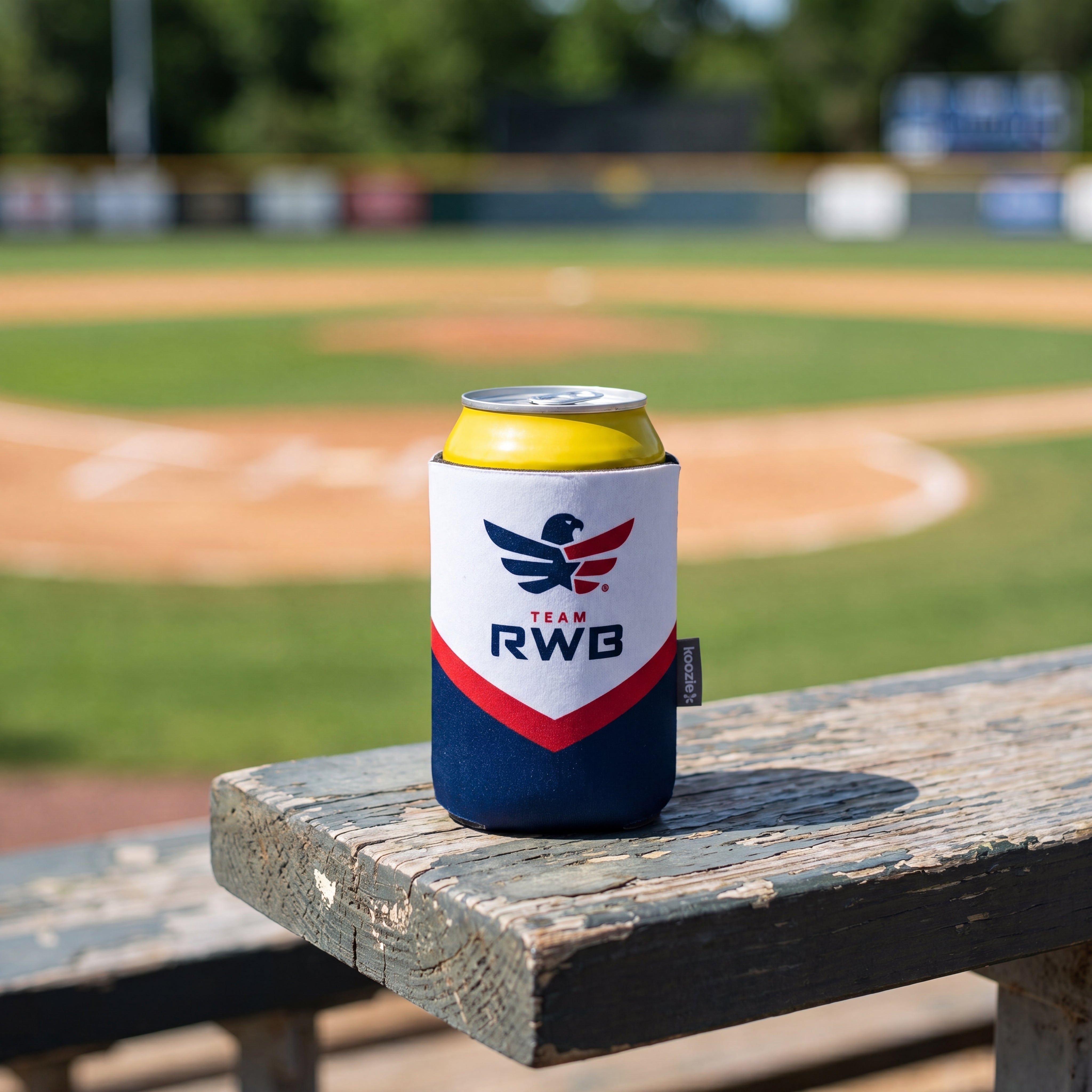 Can cooler with 'Team RWB' branding on a wooden bench at a baseball field.
