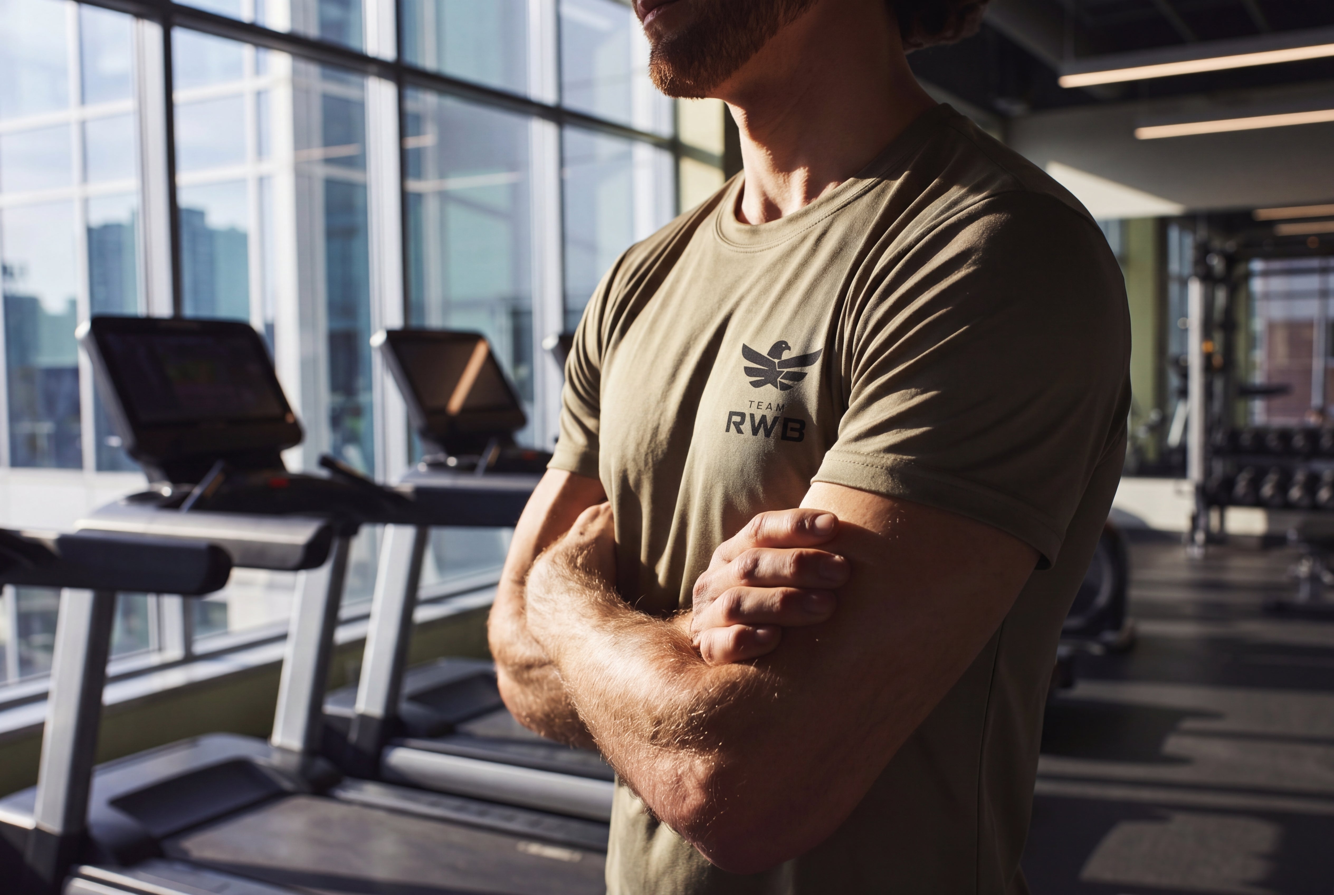 Man flexing muscles in a gym with treadmills and large windows in the background