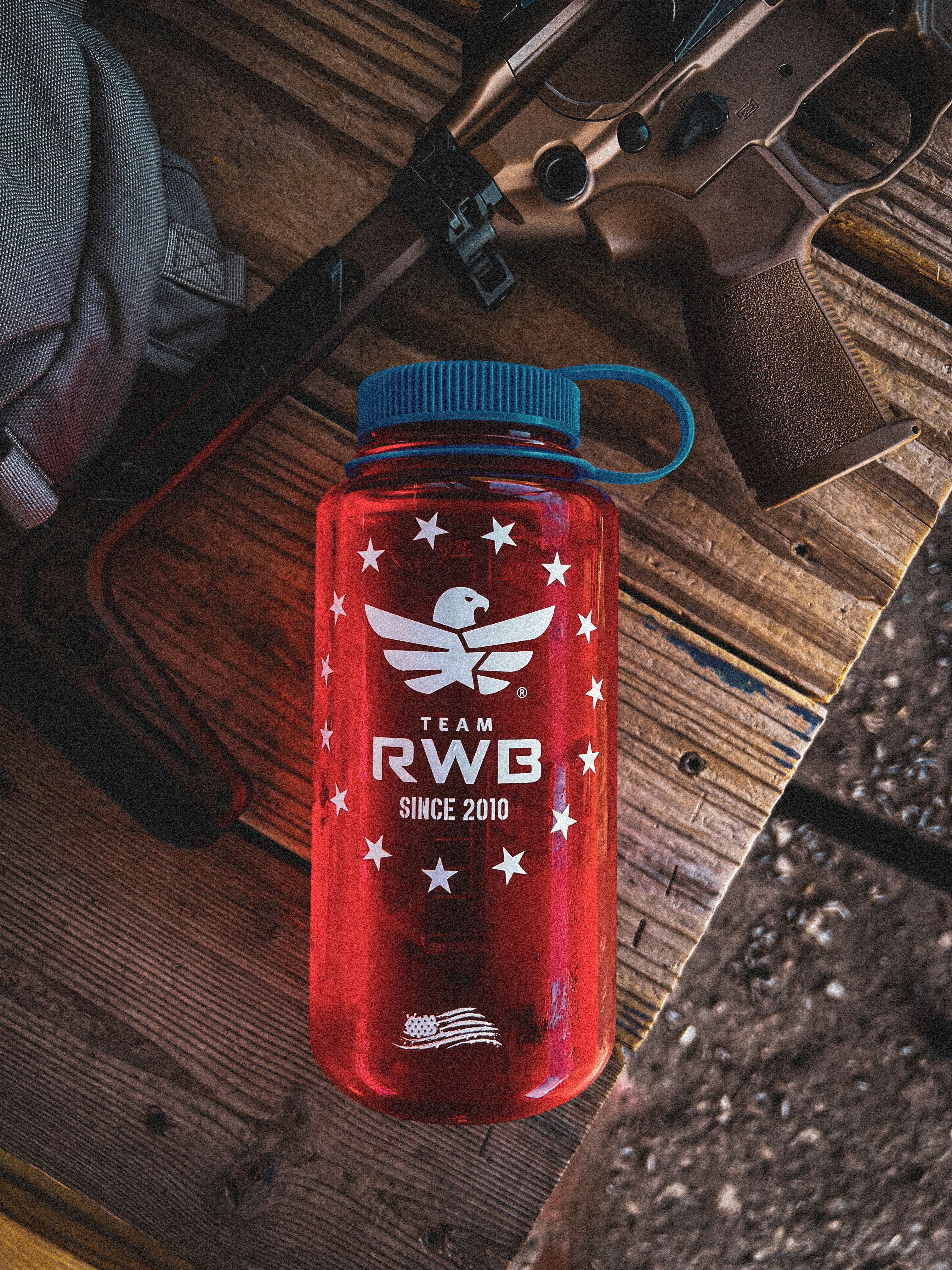 Red water bottle with blue cap featuring 'Team RWB' logo on a wooden surface with a rifle.