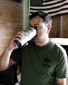 A man in an olive green Team RWB uniform undershirt t-shirt drinking from a stainless steel can, with a wooden wall and an American flag in the background. 