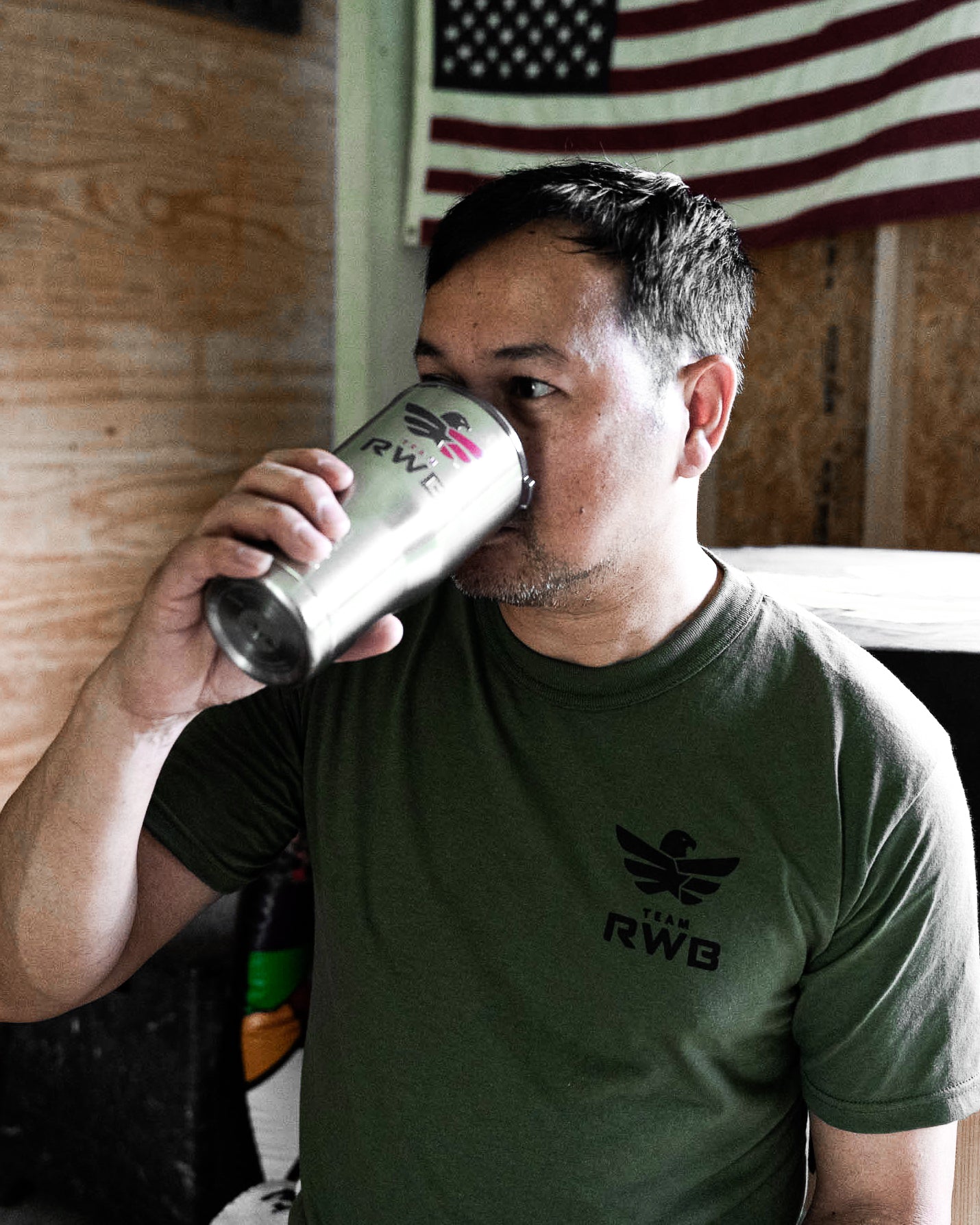 A man in an olive green Team RWB uniform undershirt t-shirt drinking from a stainless steel can, with a wooden wall and an American flag in the background. 