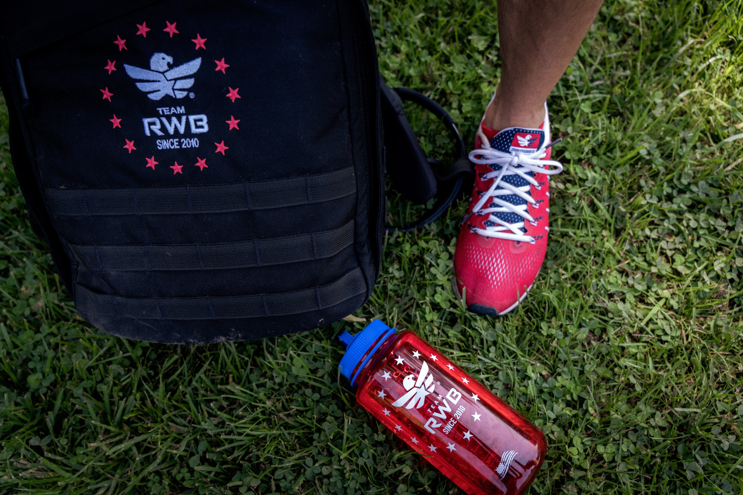 Black backpack, red running shoes, and red water bottle with 'Team RWB' logo on grass