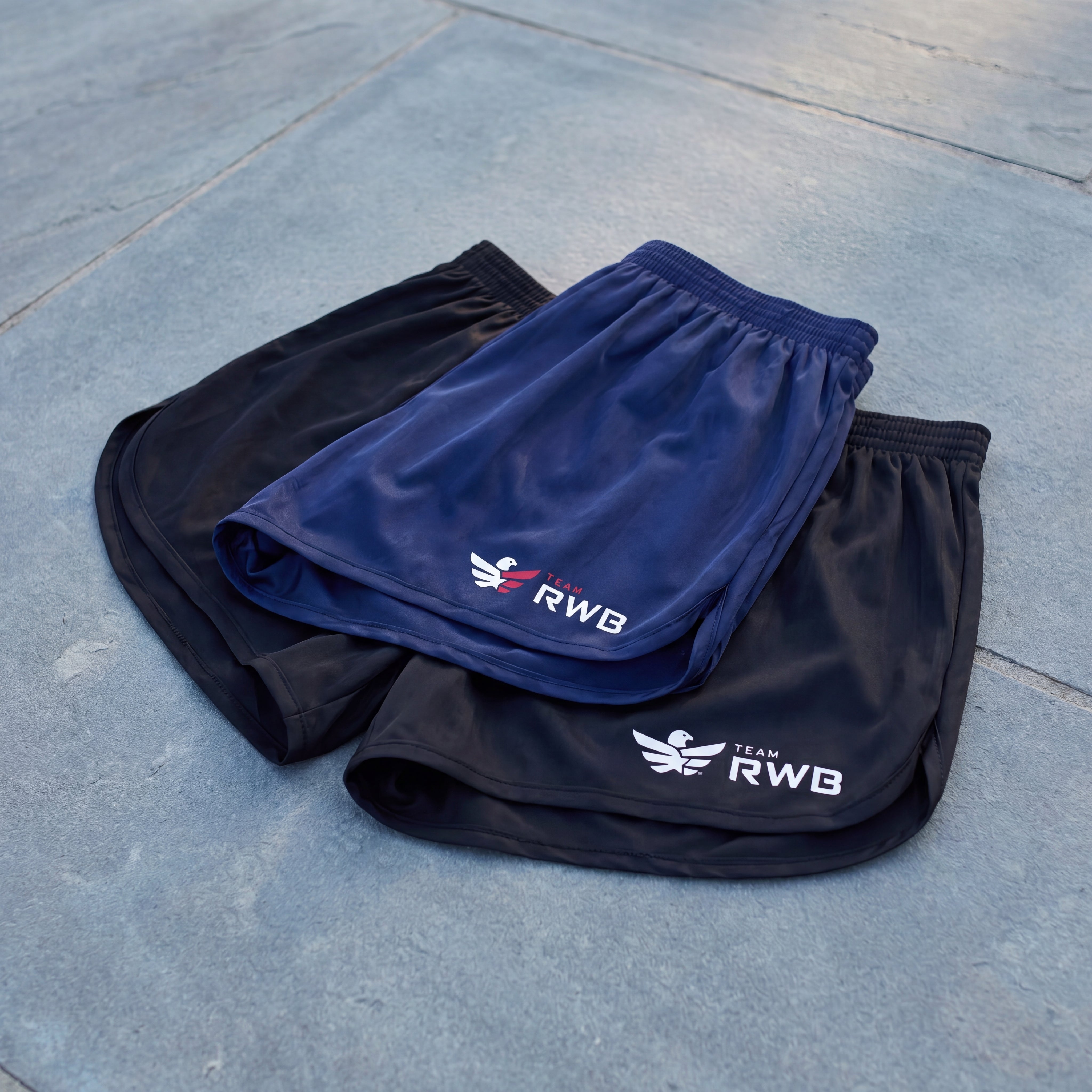 Two pairs of shorts, one blue and one black, with RWB logos on a concrete surface.